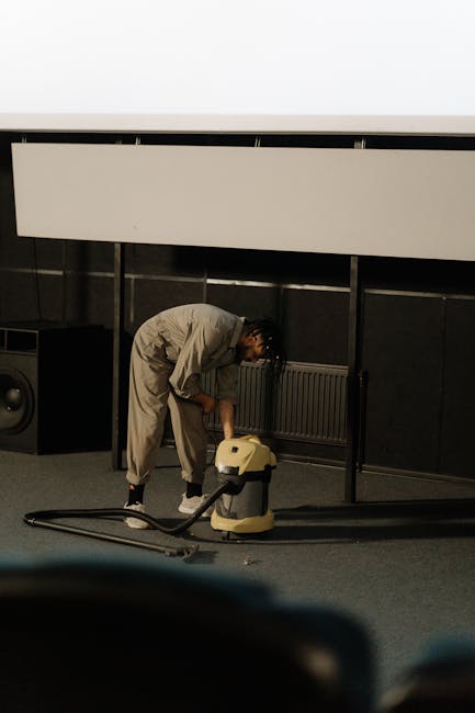 A person wearing a black leather jacket is using a vacuum cleaner with a black hose to deep clean a decorative yellow and white patterned cushion placed on a dark fabric sofa in a living room. The sce